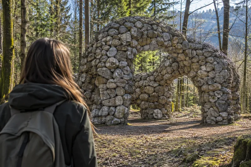 Instalación artística Simbiosi de malla metálica transparente rellena de piedras sobre una ladera boscosa.